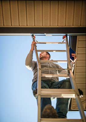 Man on ladder installing an overhead door or panel against a clear blue sky.