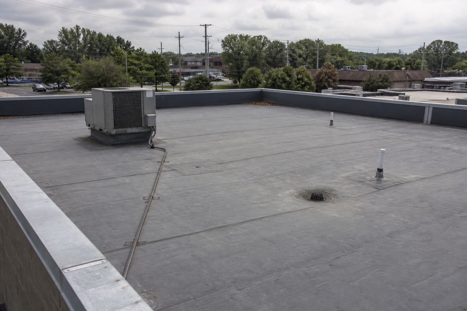 Flat commercial rooftop with vents, drains, and distant trees under cloudy skies.